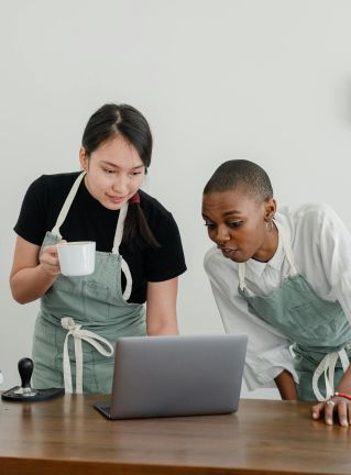 Multiethnic female baristas watching video on laptop with surprised faces while enjoying free time during coffee break at work near table with coffeemaker